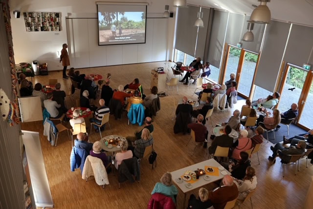 Versammlung von Menschen, die einen Film auf einer Leinwand sehen; helle Wände, Holzfußboden, entspannte Stimmung.