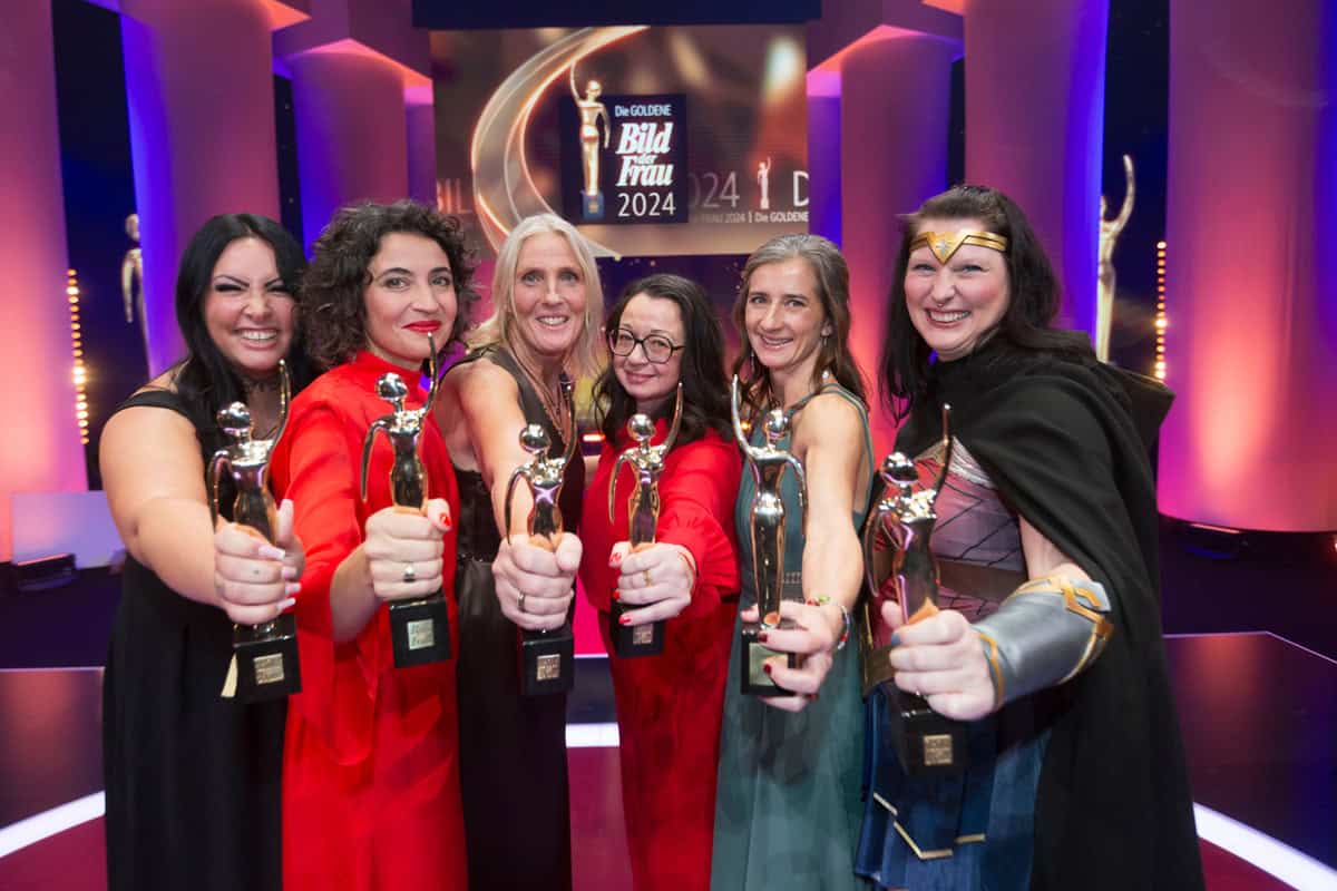 Sieben lächelnde Frauen in roten und schwarzen Outfits halten Trophäen hoch. Bühne im Hintergrund.